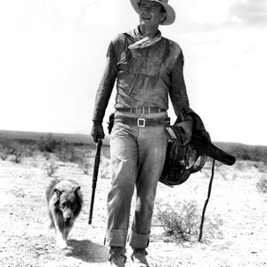 May include: A black and white photo of a cowboy wearing a hat and carrying a rifle and a saddle. He is walking with a dog in a desert landscape.