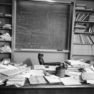 May include: A black and white photo of a cluttered desk with a chalkboard in the background. The desk is covered in papers, books, and other items. There is a chair in the background and a bookshelf on the right side of the image.