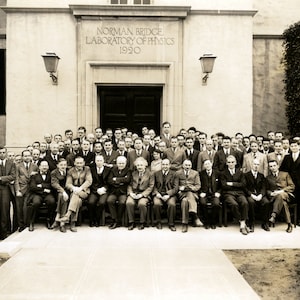 May include: A black and white photograph of a group of people standing in front of a building. The building has a sign that reads "Norman Bridge Laboratory of Physics 1920".