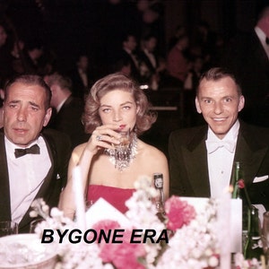 May include: A black and white photo of three people at a formal event. A man in a tuxedo is seated on the right, a woman in a red dress is seated in the middle, and a man in a tuxedo is seated on the left. The woman is holding a glass of wine and looking at the camera. The men are looking at the woman. There are flowers in the foreground.