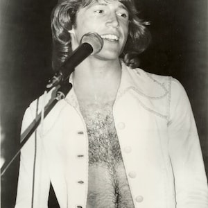 May include: A black and white photo of a man with long hair singing into a microphone. He is wearing a white shirt with buttons and his chest hair is visible.