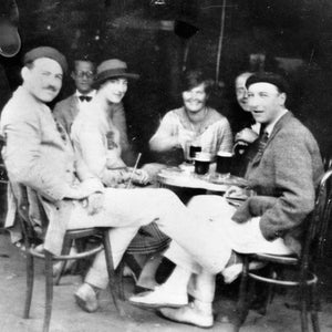 May include: Black and white photograph of a group of people sitting around a table at an outdoor cafe. The people are dressed in vintage clothing and are enjoying drinks. The image has a vintage aesthetic.