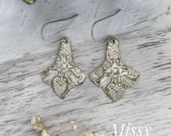 May include: A pair of silver earrings with a floral design. The earrings are hanging from a silver hook. The earrings are on a white wooden surface.
