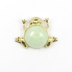 May include: A gold-toned frog brooch featuring a large, smooth, pale green cabochon stone as its body. The frog has a polished metallic finish, detailed limbs, and two small, clear, round stones set as eyes on its head, creating a distinct animal shape.