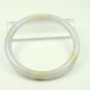 May include: A translucent jade bangle bracelet. The bracelet is a pale lavender color with subtle yellow-brown hues. The circular design is smooth and polished, showcasing the natural variations in the stone.