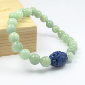 Puede incluir: Una pulsera de cuentas con cuentas redondas y lisas de color verde pálido similar al jade. La pulsera incluye una cuenta prominente de color azul oscuro, de forma ovalada, con intrincados patrones tallados y simétricos.