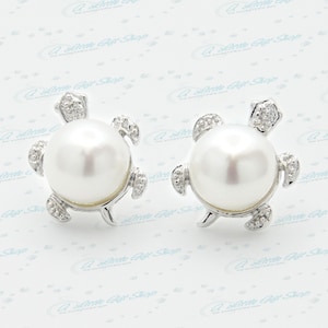 Puede incluir: Un par de pendientes de plata en forma de tortuga con una perla blanca en el centro de cada tortuga. Las tortugas tienen pequeñas piedras transparentes en su caparazón.