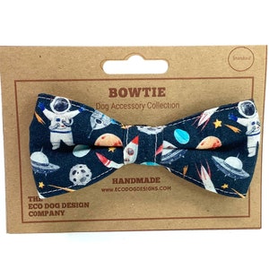 May include: A blue bow tie with a space-themed print featuring astronauts, planets, rockets, and stars. The bow tie is handmade and comes with a brown card tag that reads "BOWTIE Dog Accessory Collection" and "HANDMADE www.ECOOOGDESIGNS.COM".