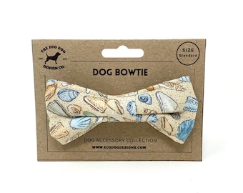 May include: A dog bowtie with a seashell pattern in beige, blue, and tan. The bowtie is on a brown cardboard backing that reads "DOG BOWTIE" and "DOG ACCESSORY COLLECTION". The size is standard.