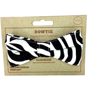 May include: A black and white zebra patterned bow tie for dogs. The bow tie is on a brown cardboard packaging with the text "BOWTIE Dog Accessory Collection" and "HANDMADE www.ecodogdesigns.com" printed on it.