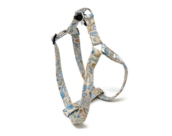 May include: A beige dog harness with a seashell pattern in blue, tan, and green. The harness has adjustable straps and metal hardware. The design features various seashell illustrations.