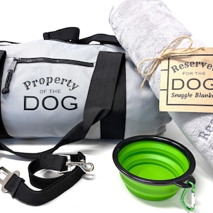 May include: A dog travel kit with a gray duffel bag, a gray and white snuggle blanket with the text "Reserved for the Dog Snuggle Blanket", a green collapsible bowl, a black leash with a clip, and a roll of green poop bags.
