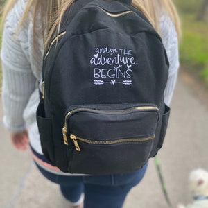 May include: A black canvas backpack with gold zips and the embroidered text "and so the adventure begins" with arrows and hearts.