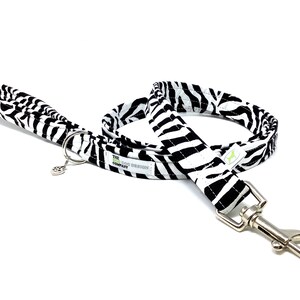 May include: A black and white zebra print dog leash with a silver clip and a green label that reads "The Dog Design Company".