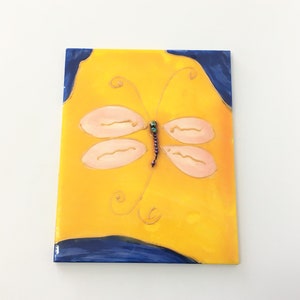 May include: A yellow ceramic tile with a blue border and a pink dragonfly painted on it. The dragonfly has a beaded body and gold accents.