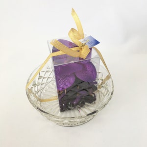 May include: A clear plastic box filled with purple orchid potpourri, tied with a gold ribbon and a price tag that reads "£30.00".