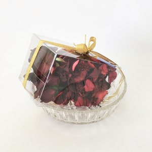 May include: A clear plastic box with a gold ribbon tied around it, filled with dried red rose petals. The box is sitting on a clear glass bowl with a decorative pattern.
