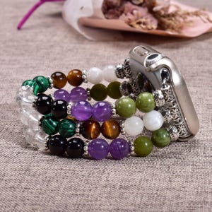 Pode incluir: Uma pulseira de relógio de vários fios com contas de pedras preciosas polidas em tons de verde, roxo, castanho, preto e branco. A pulseira está ligada a um conector de relógio de metal prateado com gravações detalhadas e acabamento liso.