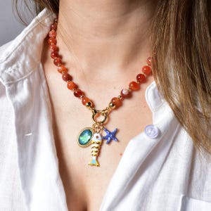 Puede incluir: Un collar de cuentas rojas y doradas. El colgante dorado tiene un dije ovalado azul y verde, un dije de pez amarillo y blanco y un dije de estrella de mar azul. El collar se usa sobre una camisa blanca.