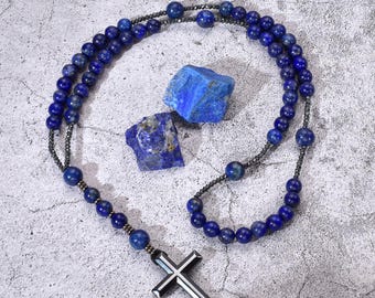 Può includere: Un rosario con perline di lapislazzuli blu scuro e un ciondolo a forma di croce argentata. Sono presenti anche due pezzi di lapislazzuli grezzi. Le perline sono infilate su un cordoncino scuro.