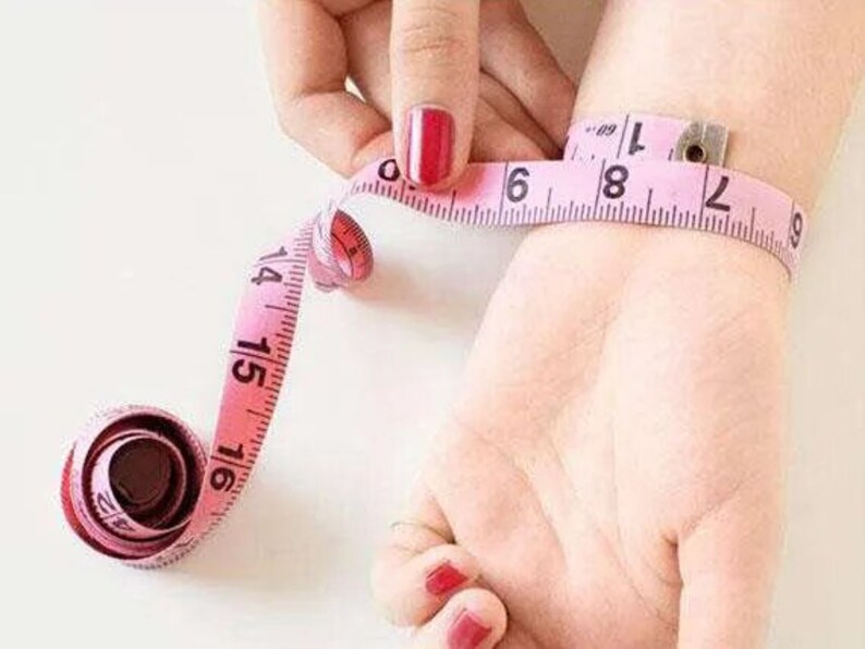 May include: A pink measuring tape wrapped around a person's wrist. The tape reads 6, 7, 8, and 9 inches.