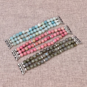 May include: Three different colored beaded Apple Watch bands. The bands are made of light blue, pink, and gray stones. Each band has a silver clasp.