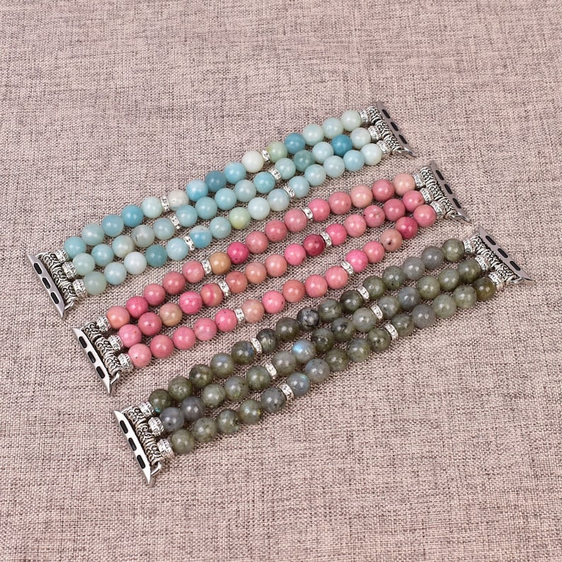 May include: Three different colored beaded Apple Watch bands. The bands are made of light blue, pink, and gray stones. Each band has a silver clasp.