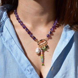 May include: A necklace with amethyst beads and gold-coloured details. It features a gold clasp and charms, including a pearl, a starfish, and a colourful fish pendant. The necklace is worn against a light blue shirt.