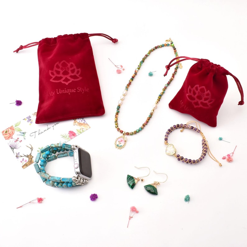 May include: A set of jewelry featuring a necklace with a floral pendant, a bracelet with purple stones, a pair of green earrings, and a turquoise and silver watch band. The jewelry is displayed on a white background with red velvet pouches and a thank you card.