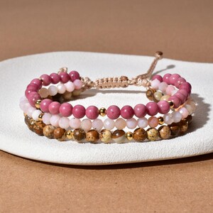 Puede incluir: Tres pulseras de cuentas con diferentes colores rosa, marrón y crema. Las pulseras están hechas de cuentas redondas y están conectadas por un cordón trenzado ajustable. Se intercalan cuentas de color dorado.