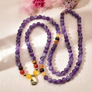 May include: Two amethyst beaded necklaces with a gold-colored pendant. The necklaces feature a variety of colorful beads, including yellow, red, brown, and orange. The pendant has a lotus flower design.