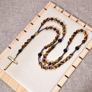 May include: A rosary with tiger eye beads and black accent beads. The rosary features a gold-colored cross and a decorative medallion. The beads are strung on a dark chain. The rosary is displayed on a white surface within a wooden frame.