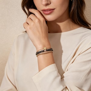 May include: Three bracelets stacked on a wrist. The top bracelet is gold with a lotus flower charm. The middle bracelet has dark grey beads and a light-coloured bead. The bottom bracelet is a gold chain.