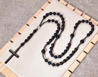 Puede incluir: Un rosario negro con un colgante de cruz y cuentas redondas. El rosario se exhibe sobre una tela de color claro, dentro de un marco de madera. La cruz tiene un diseño simple y sólido.