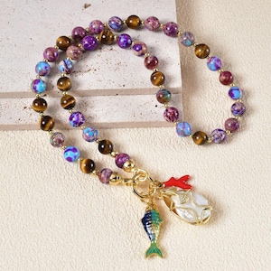 Puede incluir: Un collar de cuentas con una mezcla de cuentas moradas, azules y marrones. El collar tiene un cierre dorado con dos colgantes: un pez azul y verde y un colgante blanco en forma de perla con detalles rojos.