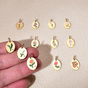 May include: Gold-coloured oval pendants featuring diverse floral designs in pink, purple, and white. Each pendant has a small loop for hanging and a tiny crystal. The pendants are shown on a neutral surface.