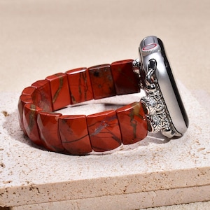 Pode incluir: Uma pulseira de relógio com contas retangulares de jaspe vermelho com veios castanhos escuros, ligada a uma caixa de relógio de metal prateado por contas espaçadoras de metal prateado texturizado com padrões intrincados e acabamento polido.