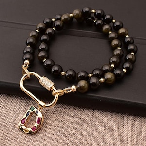 May include: A gold chain necklace with a black and gold beaded strand and a gold charm with the letter D decorated with colourful gemstones.