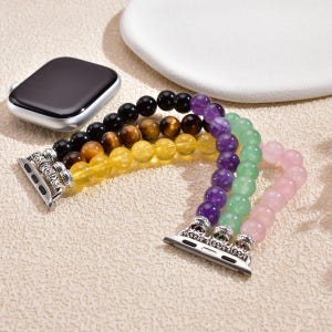 May include: A multi-coloured beaded Apple Watch strap featuring black, brown, yellow, purple, green, and pink beads. The strap has silver-coloured connectors and is next to a silver Apple Watch.