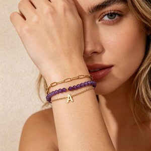 Könnte beinhalten: Drei goldene Armbänder, die an einem Handgelenk gestapelt sind. Das oberste Armband ist ein Kettenglied-Design. Das mittlere Armband hat lila Perlen. Das untere Armband hat einen kleinen goldenen Buchstaben "A"-Anhänger.