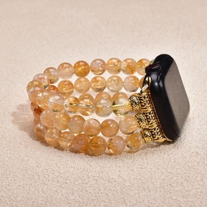 May include: A watch band featuring three strands of translucent golden rutilated quartz beads. The band connects to a black smartwatch face using three ornate, gold-toned metal cylindrical spacers with intricate engraved patterns.