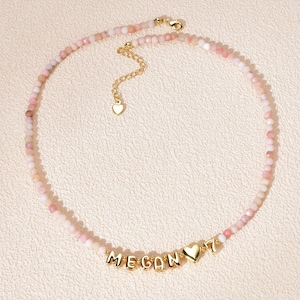 May include: A delicate necklace featuring small, light pink beads. The necklace includes gold-colored letters spelling "MEGAN", a gold heart charm, and a gold number 7 charm. The necklace has a gold chain and clasp.