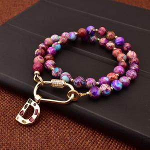 Pode incluir: Um colar dourado com um fecho e um pingente em forma da letra D. O colar é feito de contas roxas, cor-de-rosa e azuis.