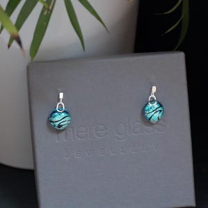 May include: A pair of silver dangle earrings with teal blue glass beads. The beads have a black and white swirl pattern. The earrings are displayed on a grey box with the text "Mere Glass Jewellery".