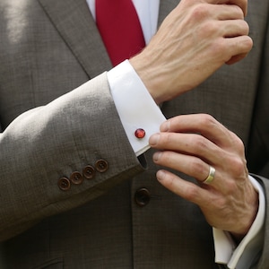 May include: A man wearing a grey suit jacket with brown buttons and a white dress shirt with cufflinks. The cufflinks are red and have a round, shiny design. The man is wearing a red tie and a silver ring on his finger.
