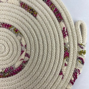 May include: A round, cream-coloured woven coaster with a pink and yellow floral pattern around the edge.