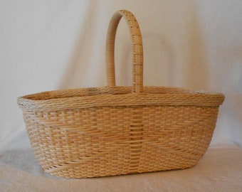 Puede incluir: Una cesta tejida natural de forma ovalada de color marrón claro con un solo asa. La cesta está hecha de finas tiras tejidas de material natural.