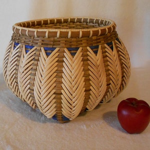 Puede incluir: Una cesta tejida con un patr&oacute;n marr&oacute;n y beige y detalles azules. La cesta tiene forma de calabaza y tiene una abertura ancha. Est&aacute; sentada sobre una superficie blanca junto a una manzana roja.