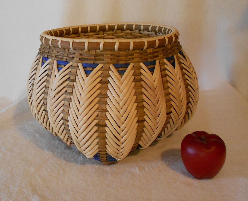Puede incluir: Una cesta tejida con un patr&oacute;n marr&oacute;n y beige y detalles azules. La cesta tiene forma de calabaza y tiene una abertura ancha. Est&aacute; sentada sobre una superficie blanca junto a una manzana roja.