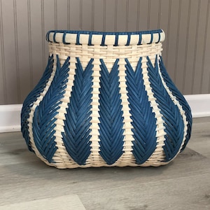 May include: A woven basket with a rounded base and a flared top. The basket features a pattern of alternating blue and natural-colored woven strips. The blue strips form a repeating chevron design, creating a decorative storage container.